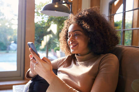 Smiling Woman Relaxing In Chair At Home Checking Social Media On Mobile Phone