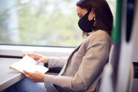 Businesswoman On Train Relaxing And Reading Book Wearing PPE Face Mask During Health Pandemic