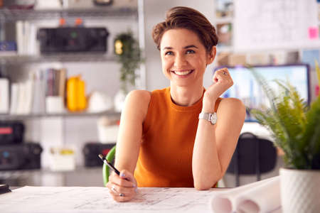 Portrait Of Female Architect In Office Working At Desk Making Notes On Plan Or Blueprintの写真素材