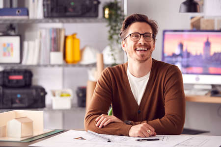 Smiling Male Architect In Office Working At Desk With Wooden Model Of Buildingの写真素材