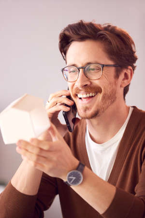 Male Architect In Office Working At Desk Taking Call On Mobile Phone And Looking At Wooden Modelの写真素材