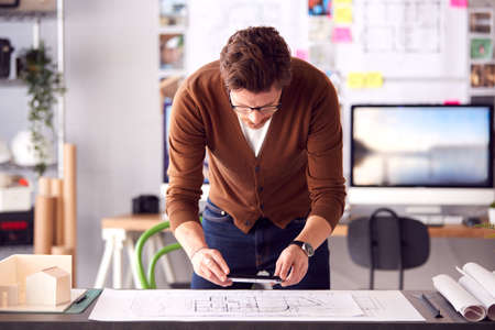 Male Architect Standing At Desk In Office Taking Photo Of Building Plans With Mobile Phone