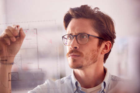 Male Architect In Office Making Notes On Building Blueprint On Transparent Screenの写真素材