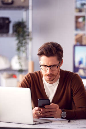 Male Architect In Office With Laptop Working On Plan At Desk Text Messaging On Mobile Phone