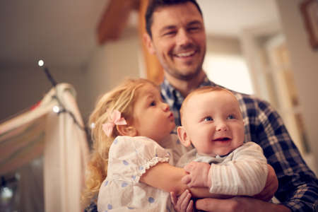 Close Up Of Young Girl Cuddling Baby Brother Whilst Sitting On Father's Knee At Home