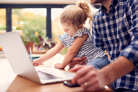 Working Father Using Laptop At Home On Kitchen Counter Whilst Looking After Young Daughterの写真素材
