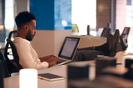 Young Businessman Working Late Sitting At Desk With Laptop In Modern Open Plan Office