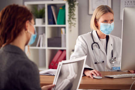 Mature Female Doctor In White Coat Wearing Face Mask Having Meeting With Woman Patient In Office