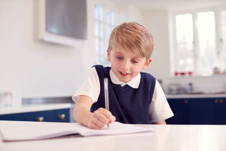 Boy At Home Wearing School Uniform Doing Homework On Kitchen Counter
