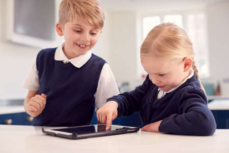 Children Wearing School Uniform In Kitchen Playing With Digital Tablet On Counterの写真素材
