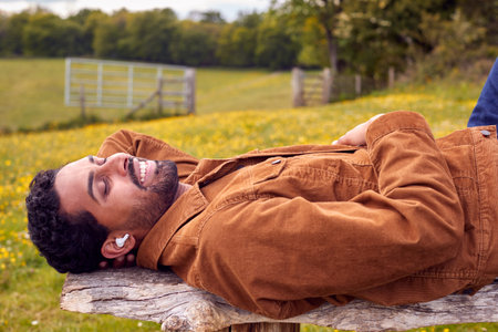 Man Lying On Bench In Countryside Relaxing And Listening To Music Or Podcast On Wireless Earphones