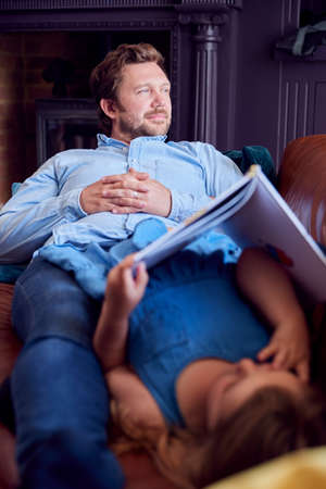 Father And Daughter Lying On Sofa At Home As Girl Reads Bookの写真素材