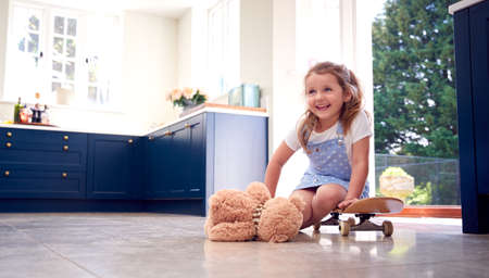 Smiling Young Girl Playing Game Sitting On Skateboard With Teddy Bear At Homeの写真素材