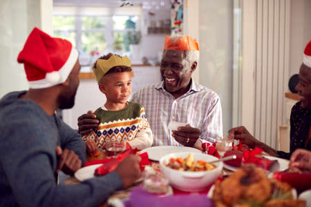 Multi Generation Family In Paper Hats Reading Christmas Cracker Jokes Before Eating Meal Together