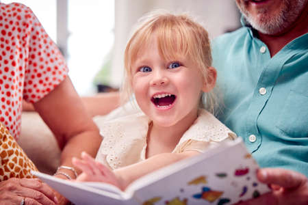 Close Up Of Grandparents Sitting On Sofa With Granddaughter At Home Reading Book Togetherの写真素材