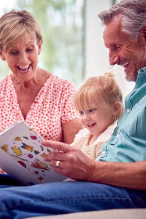 Grandparents Sitting On Sofa With Granddaughter At Home Reading Book Together