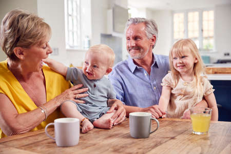 Grandparents Looking After Laughing Granddaughter And Baby Grandson Sitting At Kitchen Table