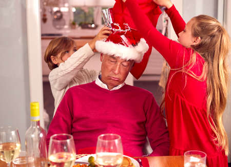 Grandchildren Having Fun With Grandfather Sleeping After Christmas Lunch