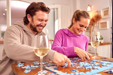 Couple Sitting At Table At Home With Glass Of Wine Doing Jigsaw Puzzle Togetherの写真素材