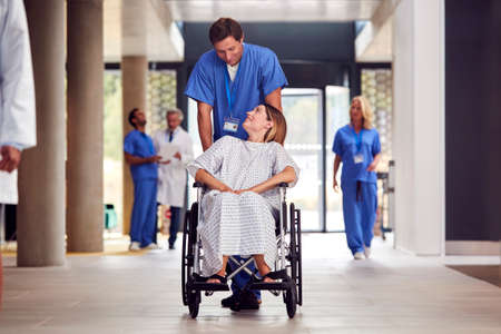 Male Nurse Wearing Scrubs Pushing Female Patient In Wheelchair Through Hospital Buildingの写真素材