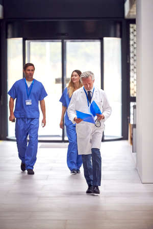 Male Doctor Wearing White Coat Reading Patient Notes Walking In Busy Hospital