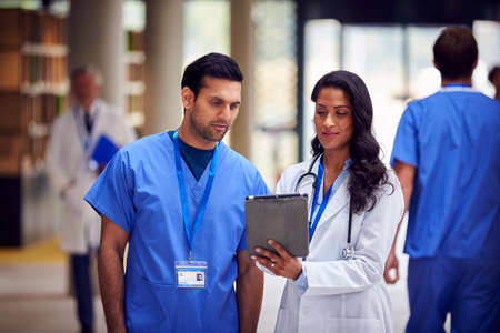 Two Medical Staff In White Coats And Scrubs With Digital Tablet Having Informal Meeting In Hospital