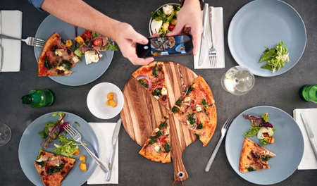 Overhead View Of Friends In Pizza Restaurant Taking Picture Of Food On Mobile Phone