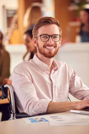 Portrait Of Businessman In Wheelchair Sitting At Desk Working On Laptop In Busy Officeの写真素材