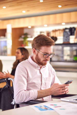 Businessman In Wheelchair With Mobile Phone Sitting At Desk Working On Laptop In Busy Officeの写真素材