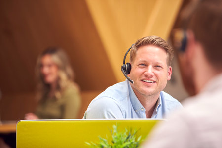 Businessman In Cubicle Wearing Headset Talking To Colleague In Busy Customer Services Centre