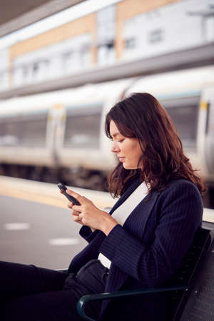 Businesswoman Commuting To Work Waiting For Train On Station Platform Looking At Mobile Phonesの写真素材