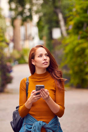 Female Vlogger Or Social Influencer Walking Through City Street Looking At Mobile Phone