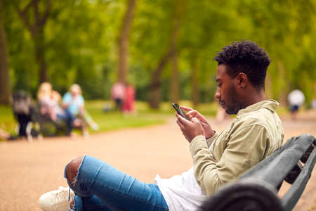 Male Vlogger Or Social Influencer Sitting On Bench In City Park Looking At Mobile Phoneの写真素材