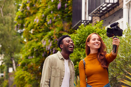 Young Couple Travelling Through City Together Vlogging To Video Camera On Handheld Tripod