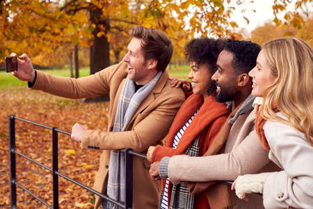 Group Of Friends Outdoors Wearing Coats And Scarves Posing For Selfie On Phone In Autumn Park