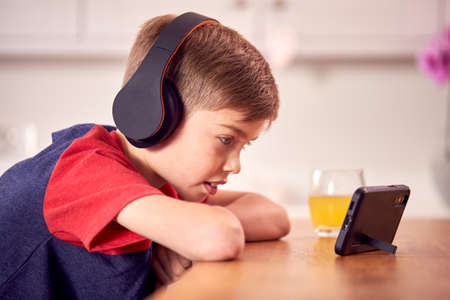 Boy At Home In Kitchen Watching Movie Or Show On Mobile Phone Wearing Wireless Headphonesの写真素材
