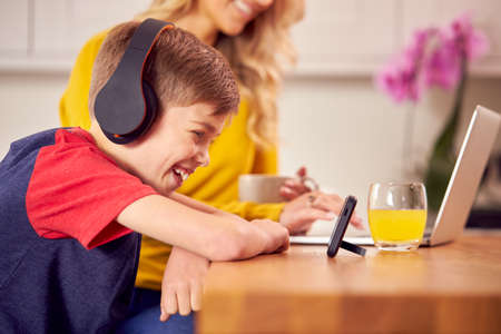 Boy In Kitchen Watching Movie On Mobile Phone Wearing Wireless Headphones As Mother Works On Laptopの写真素材