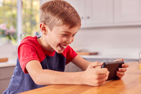 Excited Boy At Home In Kitchen Playing Game On Mobile Phone