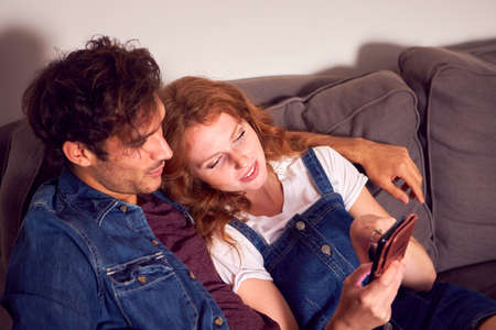 Couple Sitting On Sofa In Lounge At Home Looking At Their Mobile Phone Togetherの写真素材