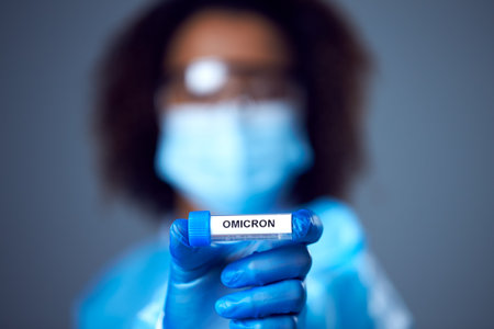 Female Lab Worker in Lab Coat And Face Mask Researching Omicron Variant Of Covid-19 Holds Test Tubeの写真素材