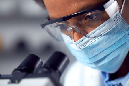 Close Up Of Female Lab Worker Wearing PPE And Safety Glasses Looking Through Microscope