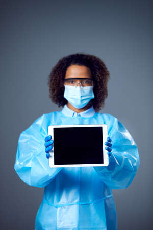 Studio Portrait Of Female Lab Worker in PPE With Face Mask And Safety Glasses Holding Digital Tablet