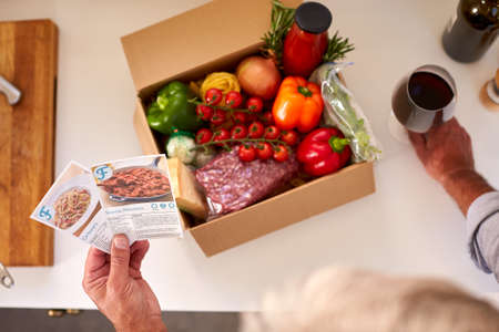 Overhead Of Man In Kitchen Holding Recipe Cards For Online Meal Food Recipe Kit Delivered To Home