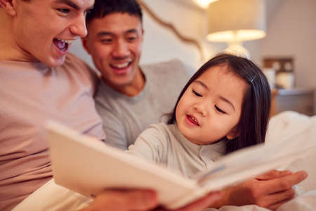 Family With Two Dads In Bed At Home Reading Story To Daughterの写真素材