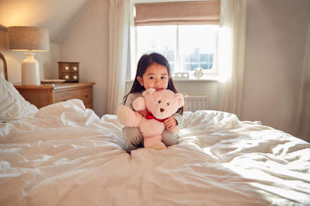 Young Girl Sitting On Bed Wearing Pyjamas At Home Cuddling Soft Toyの写真素材