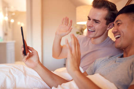 Same Sex Male Couple Lying In Bed At Home Making Video Call On Mobile Phoneの写真素材