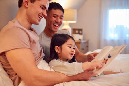 Family With Two Dads In Bed At Home Reading Story To Daughterの写真素材