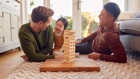 Family With Two Dads Playing Game With Daughter At Home Stacking Wooden Bricks Into Towerの写真素材