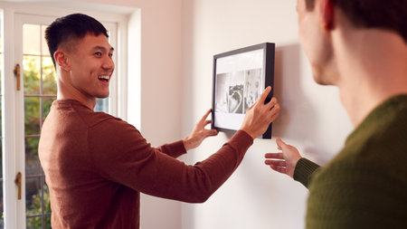 Same Sex Male Couple Hanging Picture On Wall At Home Togetherの写真素材