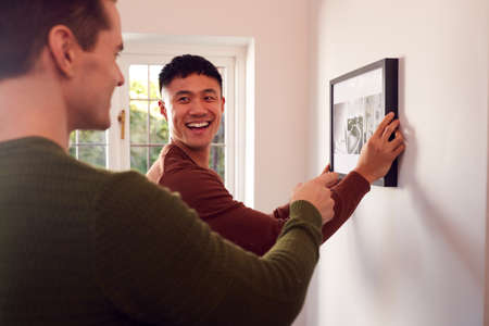 Same Sex Male Couple Hanging Picture On Wall At Home Togetherの写真素材